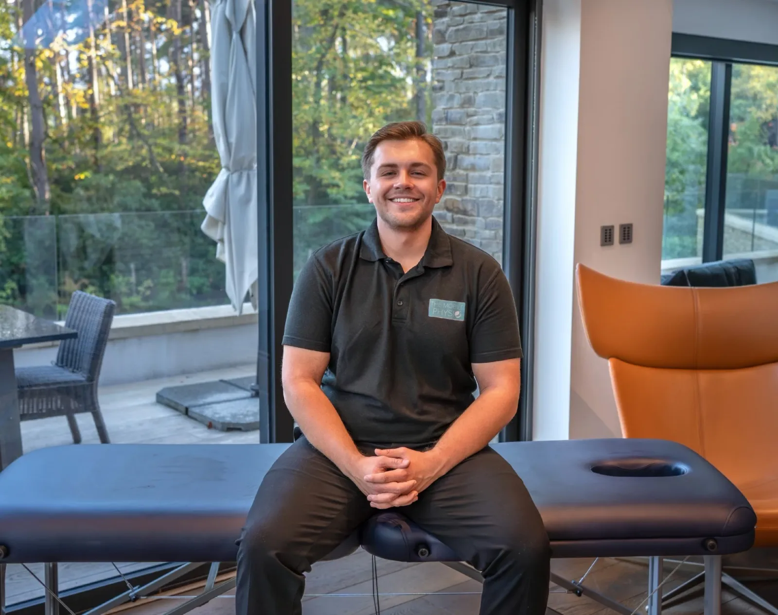 Max Gaffney, founder and physiotherapist at The Mobile Physio, smiling and seated on a portable treatment couch set up in a living room.