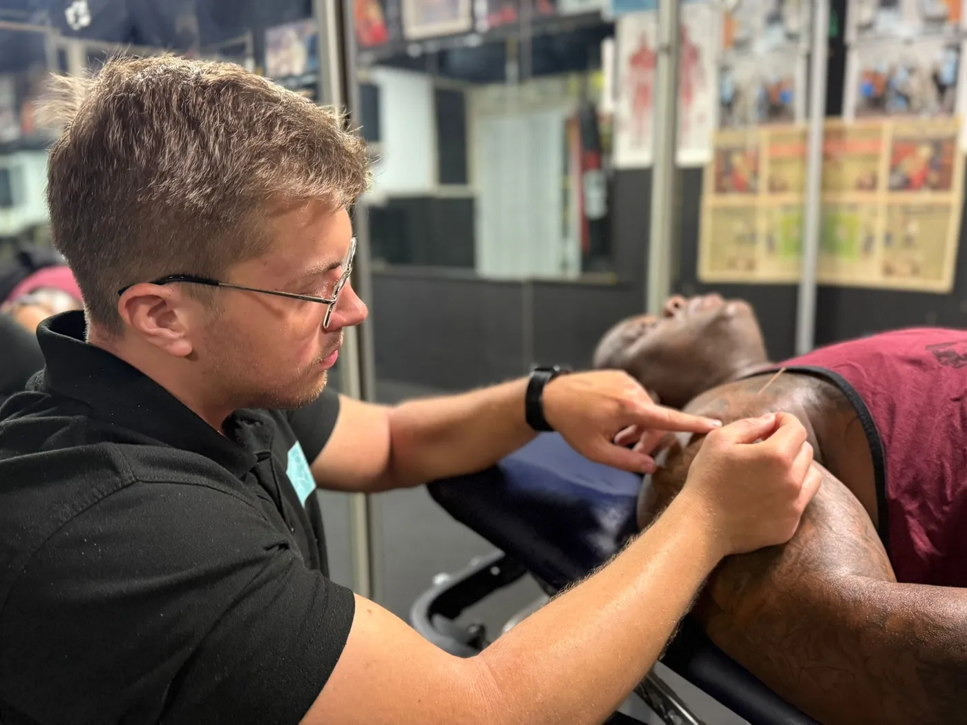 Max treating a patient lying on a treatment couch at a boxing gym, carefully placing an acupuncture needle in his shoulder.