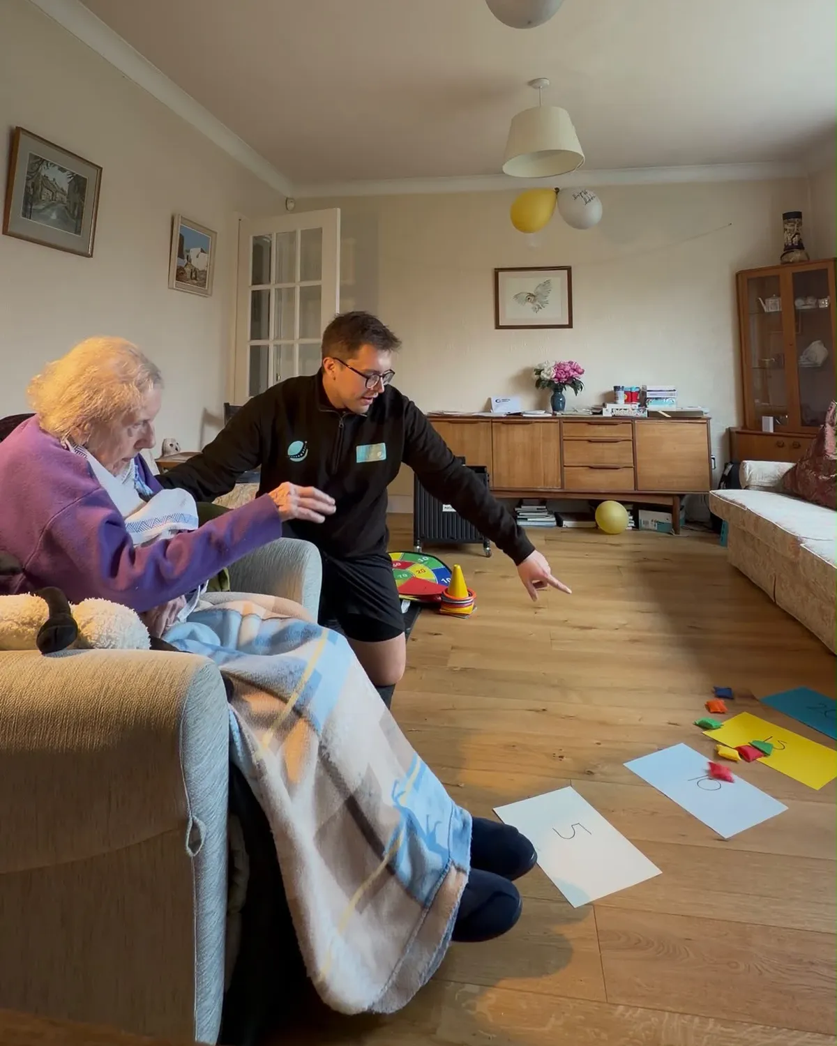Max kneeling beside an older patient seated in her armchair at home, pointing to numbered targets on the floor during a beanbag game.