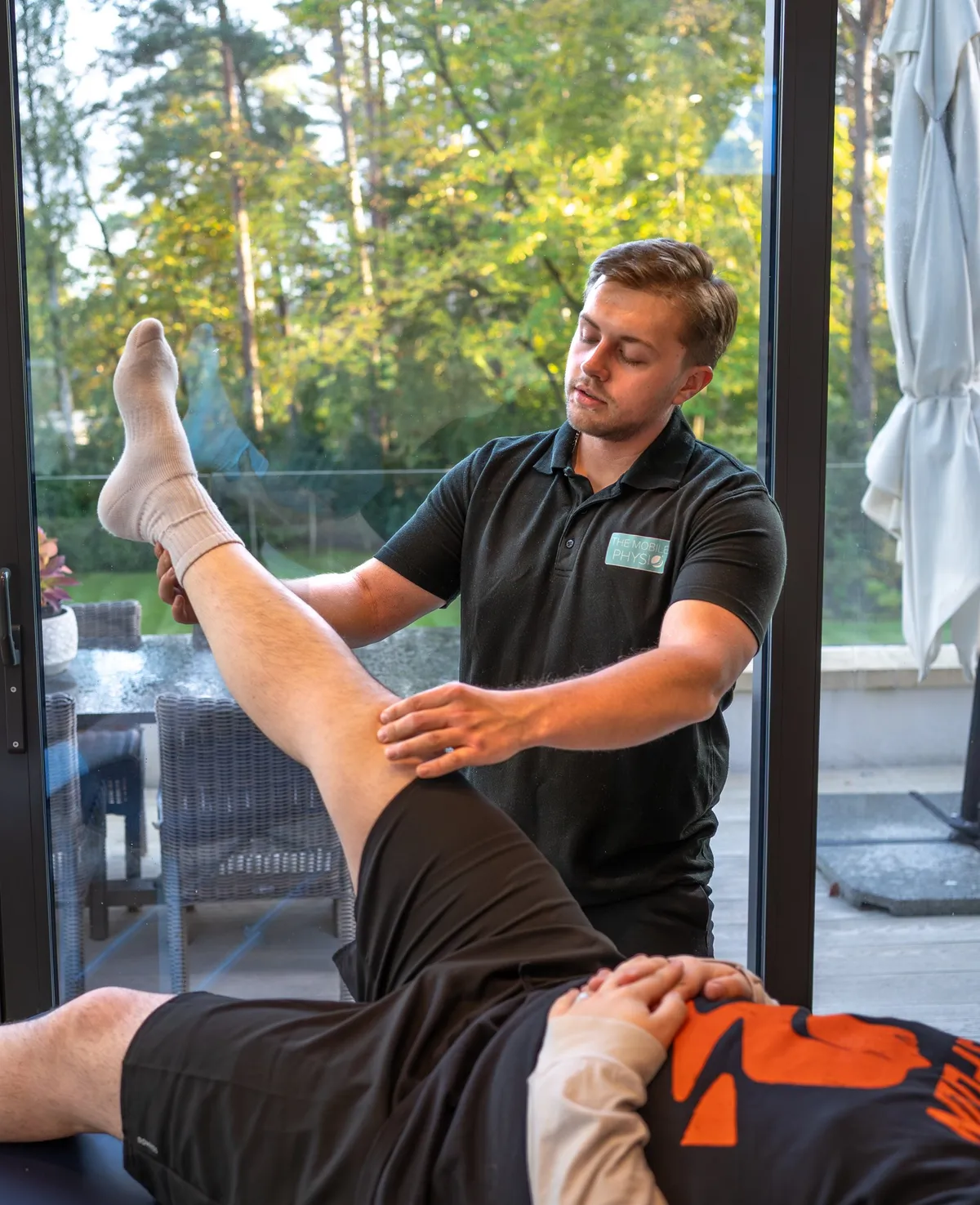 Max at a patient’s home, supporting their raised leg on a treatment couch while assessing movement at the hip and knee.