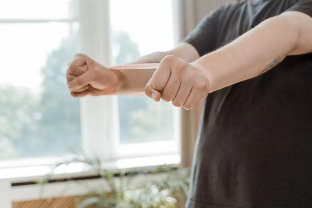 A person stretching a resistance band between their wrists beside a bright window at home.
