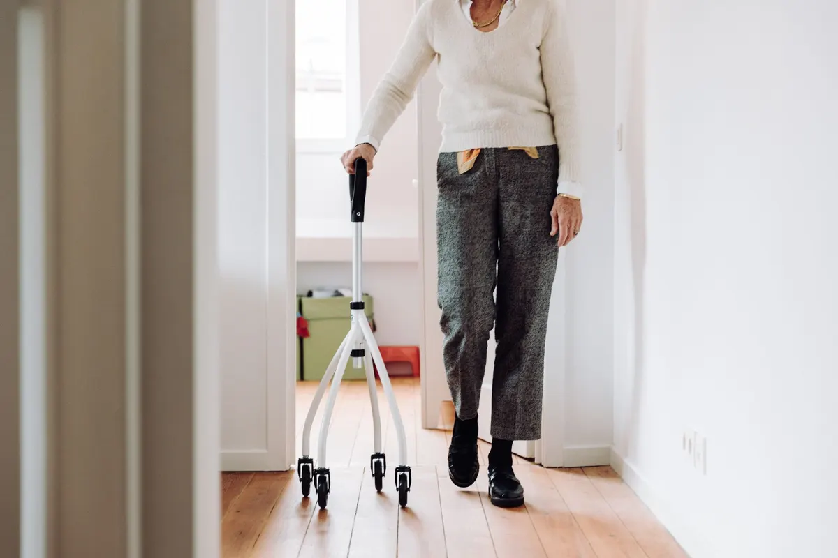 A person walking along a bright hallway at home, steadying themselves with a wheeled walking aid.